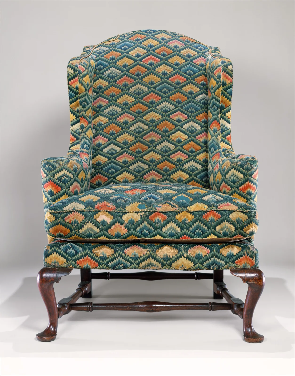 A wing-back easy chair covered in multicoloured flame-stitch needlework, standing on cabriole legs joined by turned stretchers