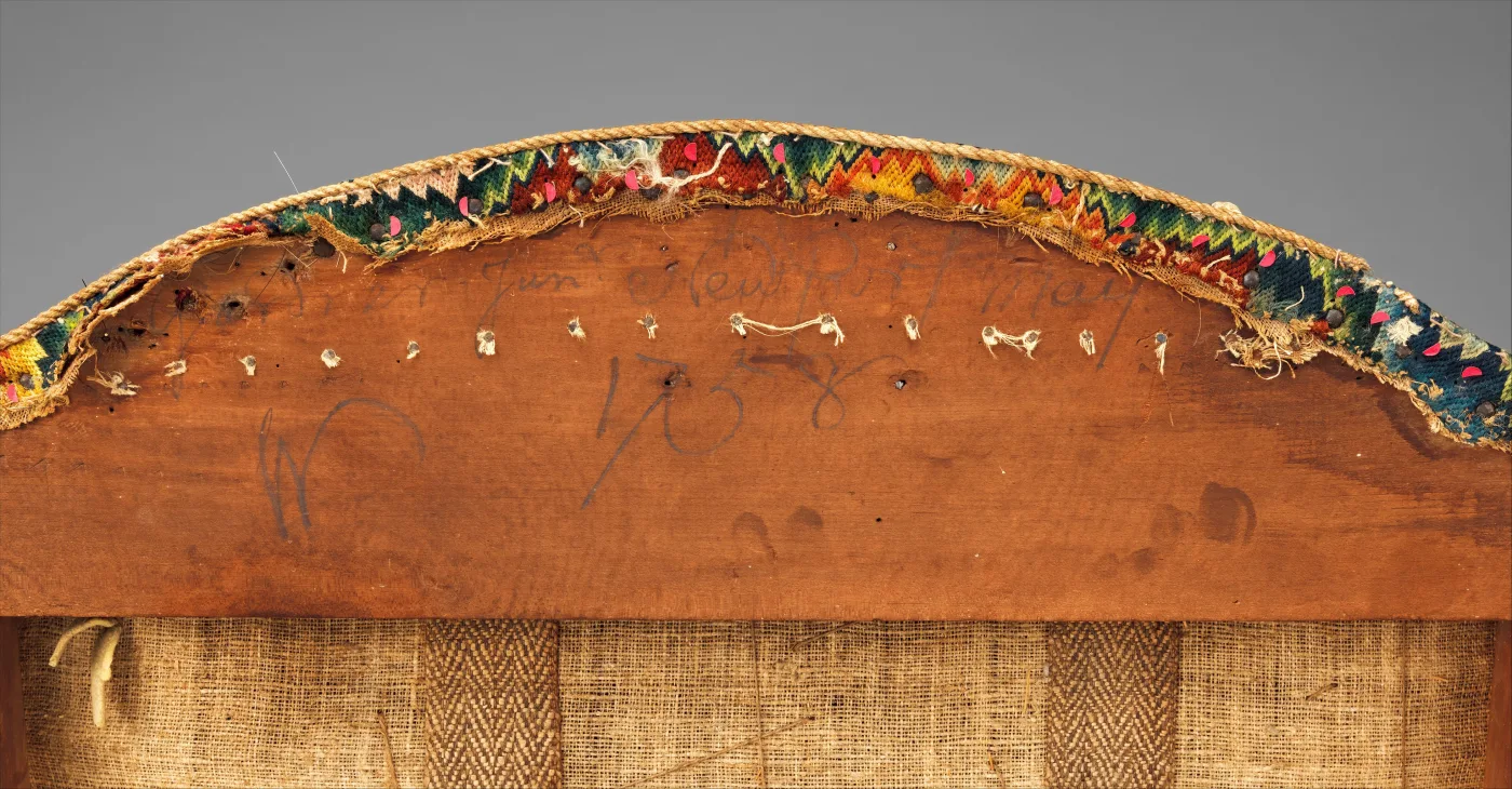 Close view of the back of a wooden chair crest rail with ink writing and the date 1758, a row of small tack holes with threads, frayed needlework along the top edge and herringbone webbing below