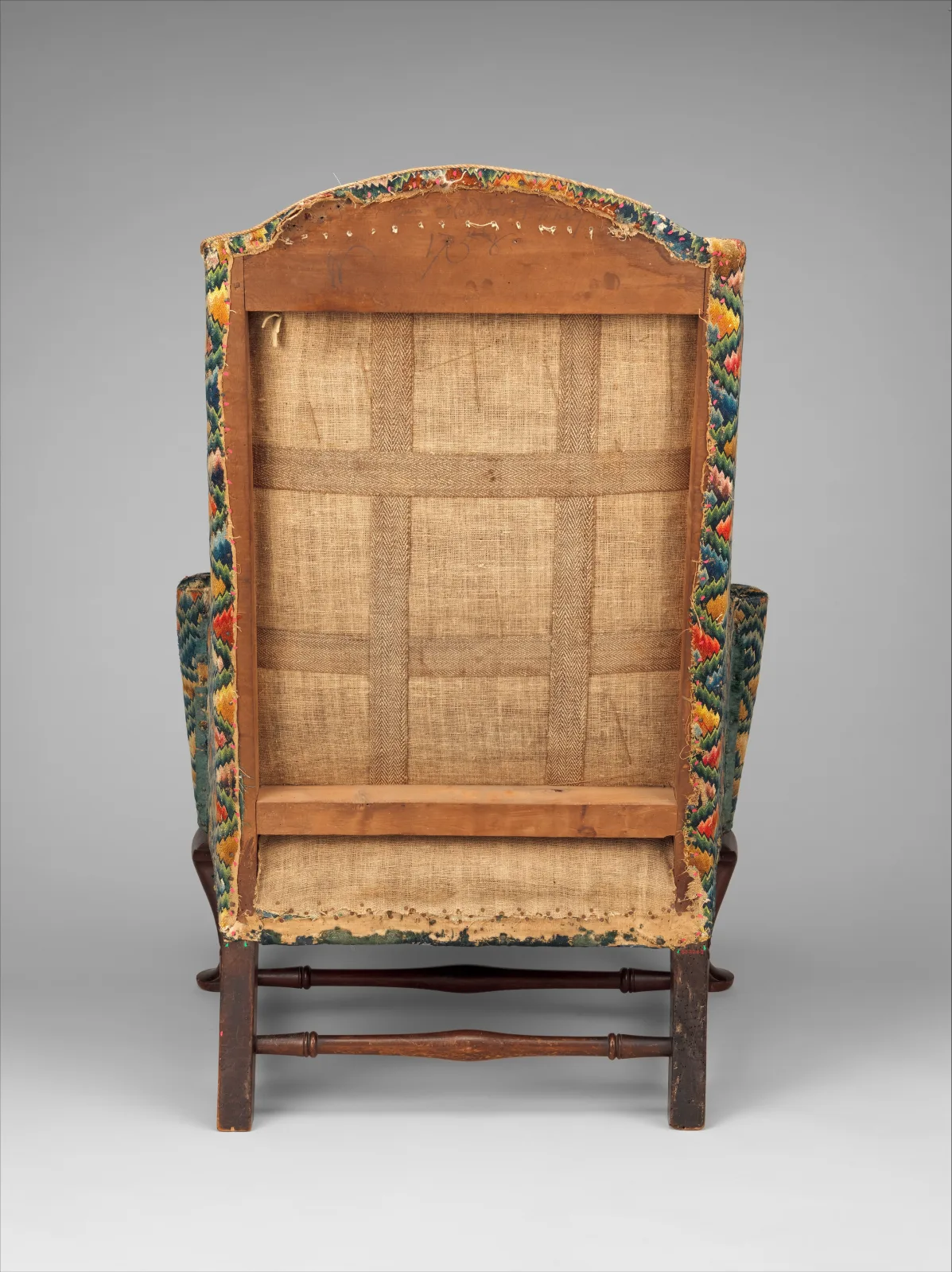 The back of a wing chair with the outer cover off, showing a grid of vertical and horizontal webbing strips over coarse linen within the wooden back frame