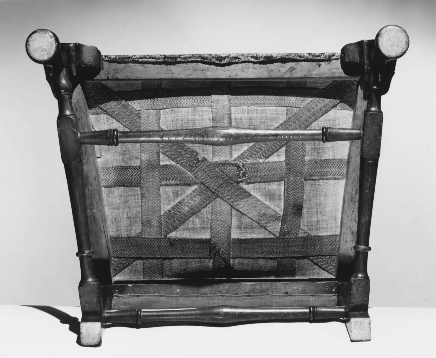 Black and white view of a chair turned upside down, showing webbing strips interlaced across the underside of the seat frame between turned stretchers