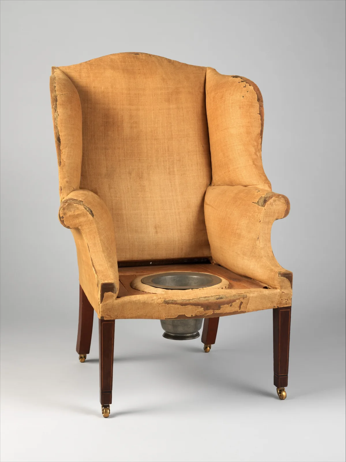 A wing easy chair covered only in worn tan muslin, with a round opening in the seat holding a pewter pan, on square tapered mahogany legs