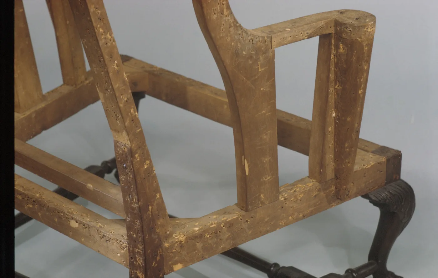 Close view of the bare wooden frame of an easy chair at the arm and seat rail, the pale wood densely marked with small dark tack holes
