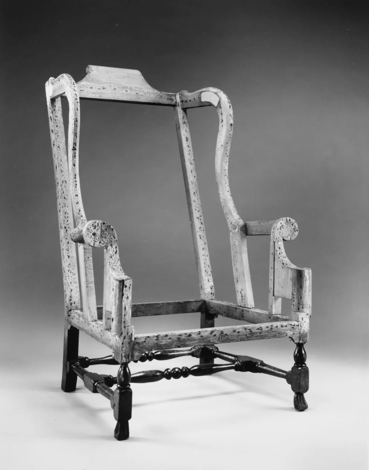 Black and white photograph of a bare wing chair frame with scrolled arms and turned legs, its pale wooden rails covered in small dark tack holes