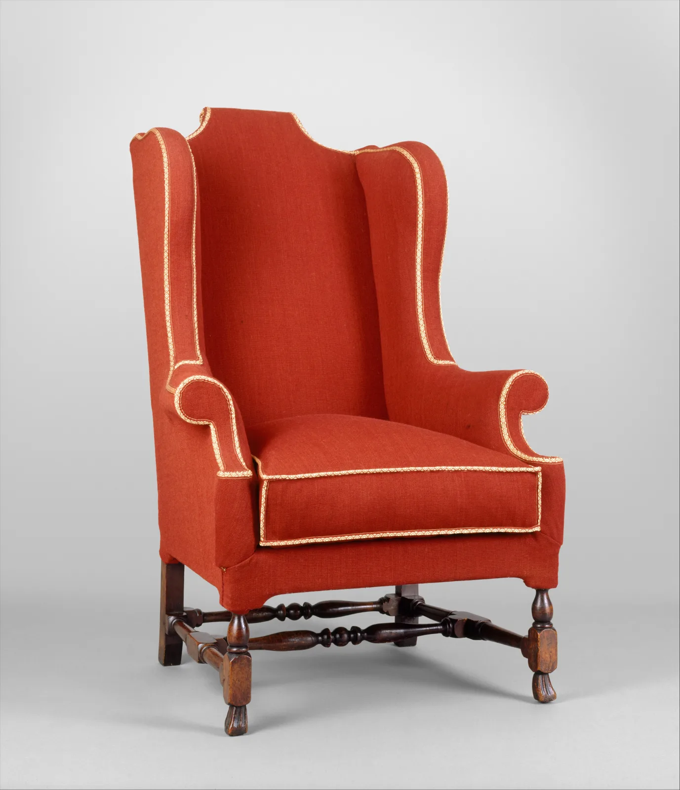 The same wing chair upholstered in red fabric with cream welting along the wings, arms and cushion, on turned legs and stretchers