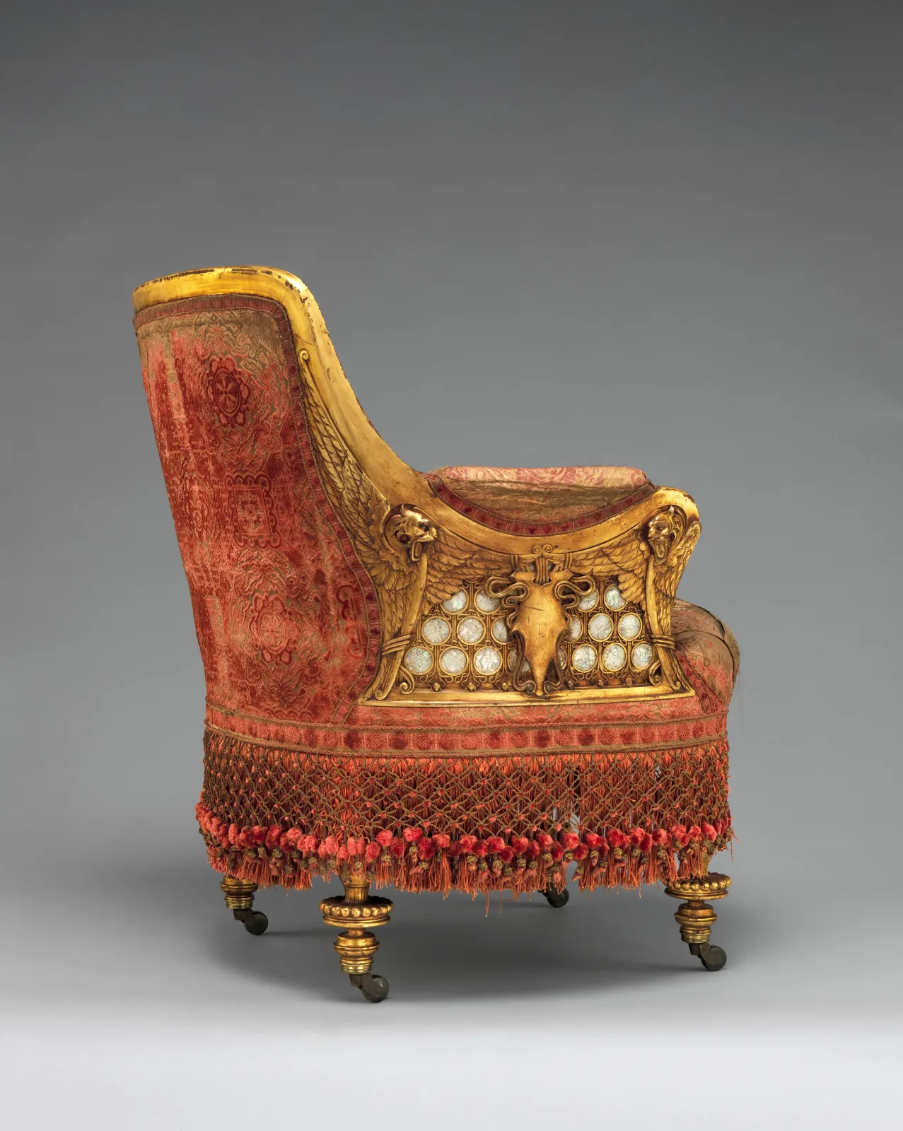 A low gilded armchair upholstered in faded red patterned silk velvet with a deep red fringe around the seat, mother-of-pearl discs set in the arm panel, on small turned feet with casters