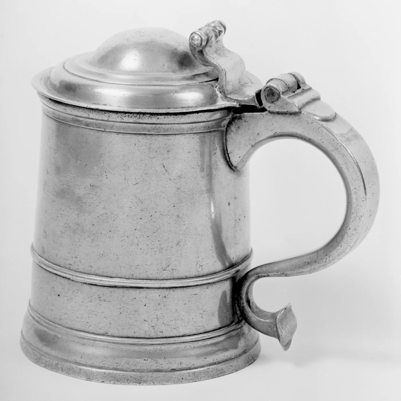 Straight-sided pewter tankard with a domed lid, a heavy scrolled handle and moulded bands around the body, photographed in black and white