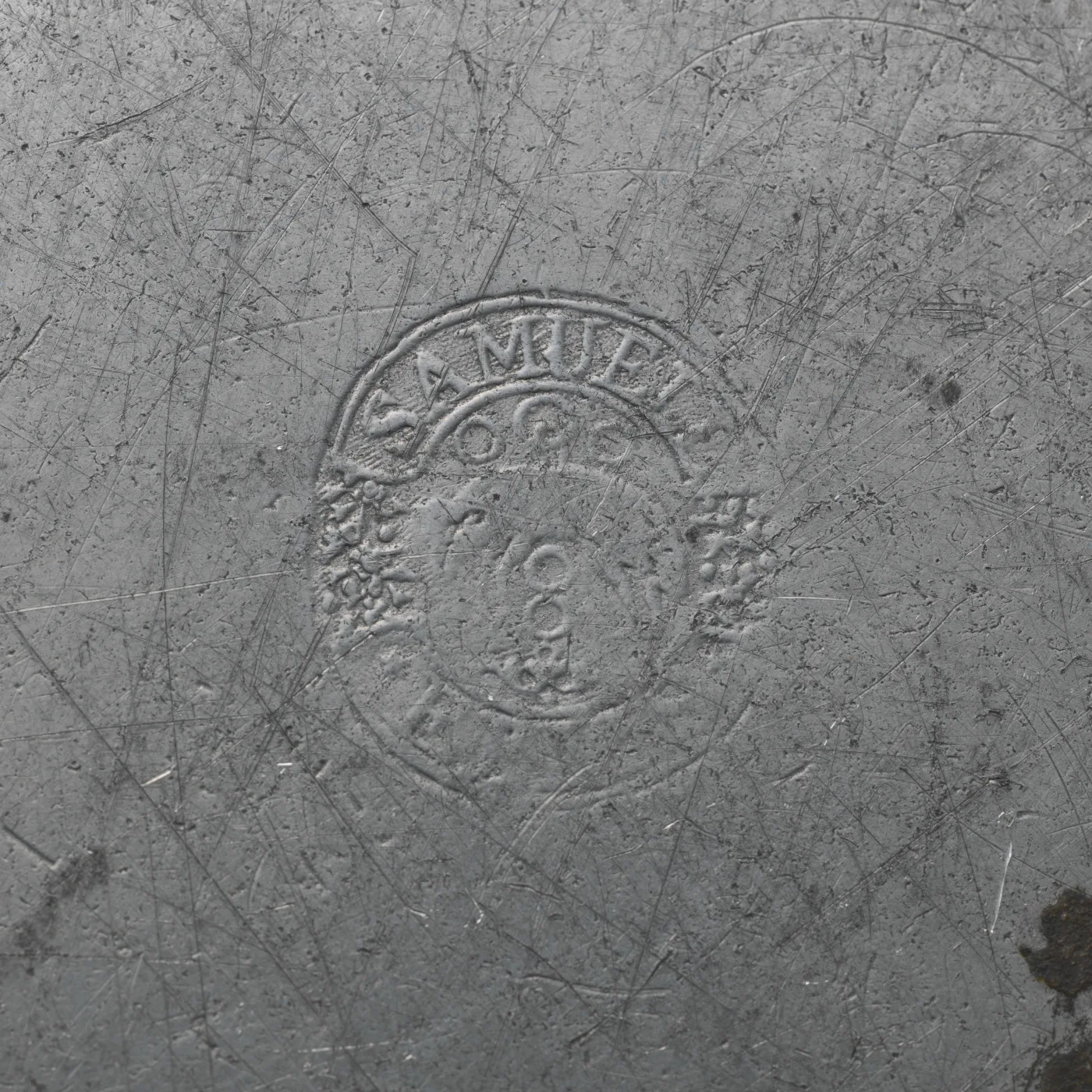 Close view of a grey pewter surface covered in fine criss-cross knife scratches, with a circular struck touch reading SAMUEL ELLIS around a central device