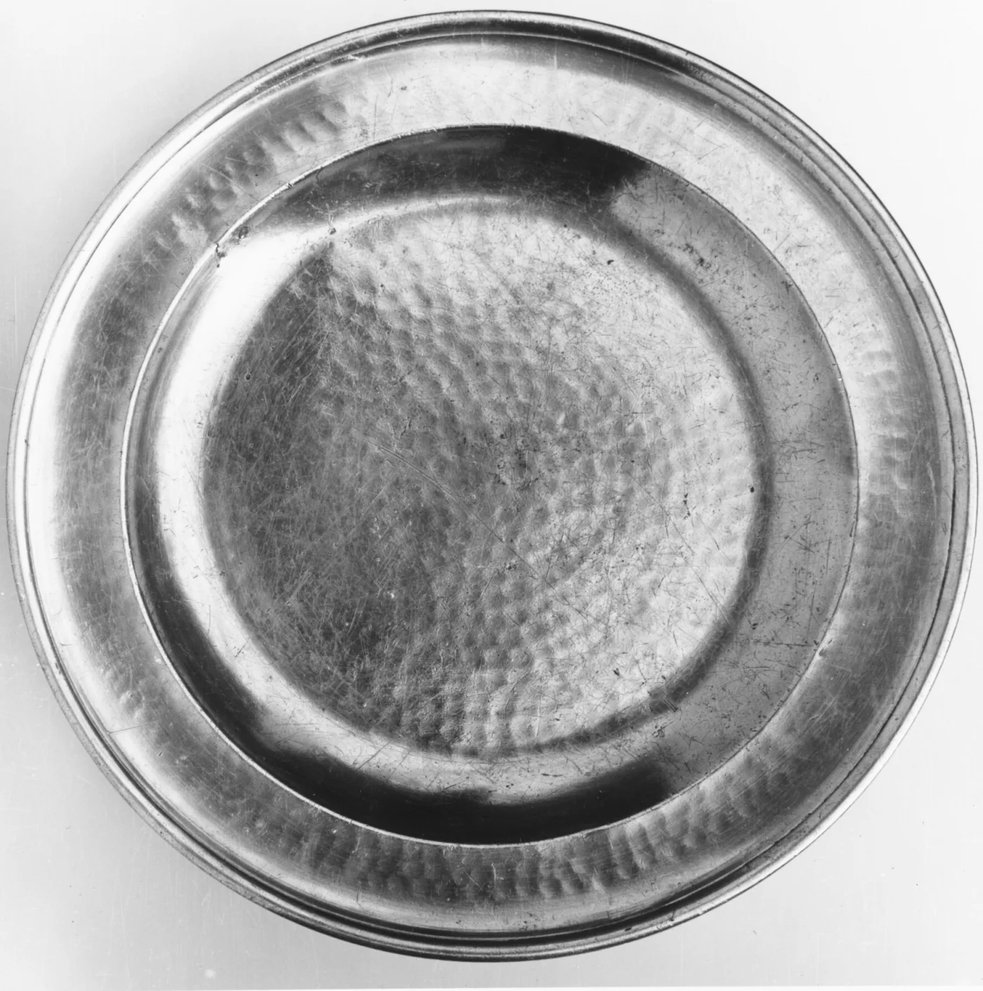 Round pewter dish seen from above with rows of small hammer dents across the curved wall and into the centre