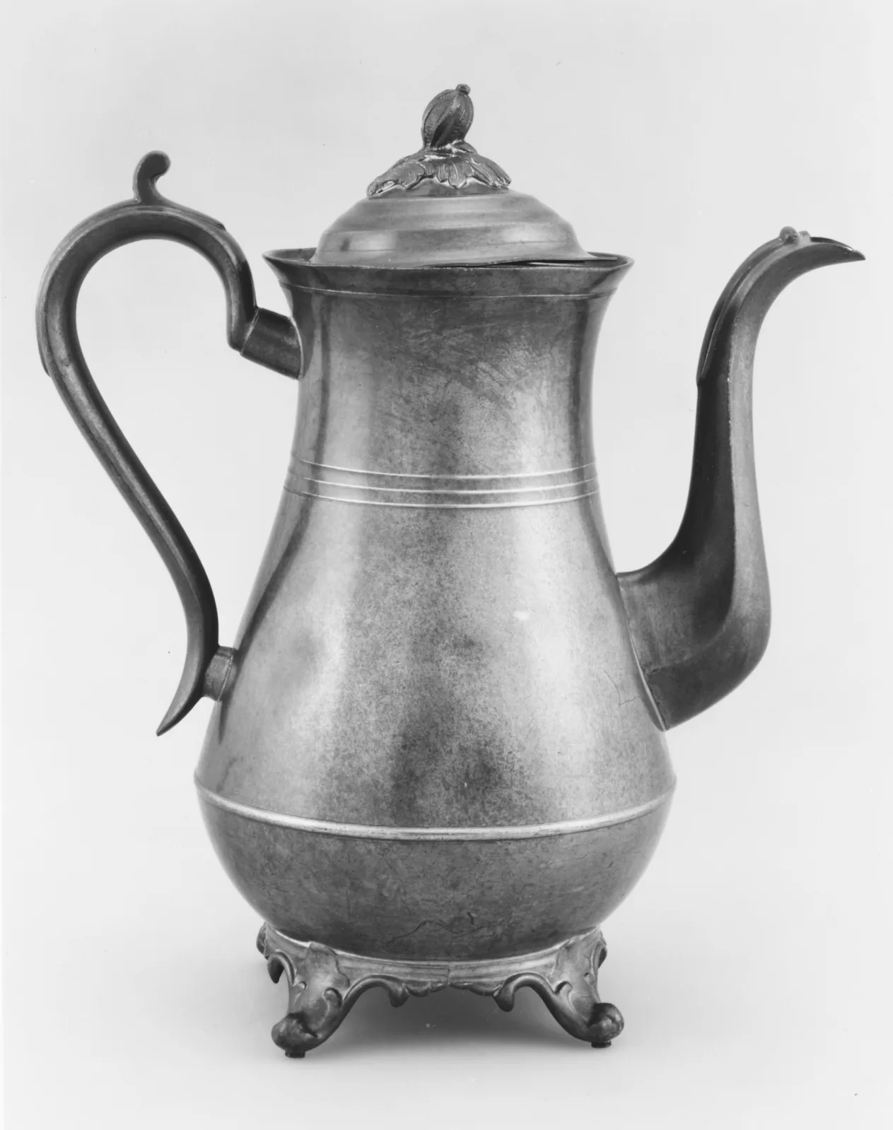 Tall pear-shaped coffeepot in grey Britannia metal with a scrolled handle, a long curved spout, incised bands and four scrolled feet, photographed in black and white