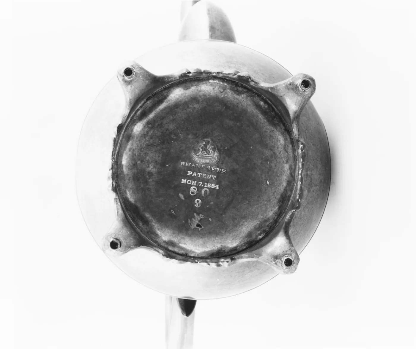 Underside of a coffeepot showing a round base with four scrolled feet attached by rough solder joins, and stamped lines reading R.W. ANDREWS PATENT MCH. 7 1854 and two numbers