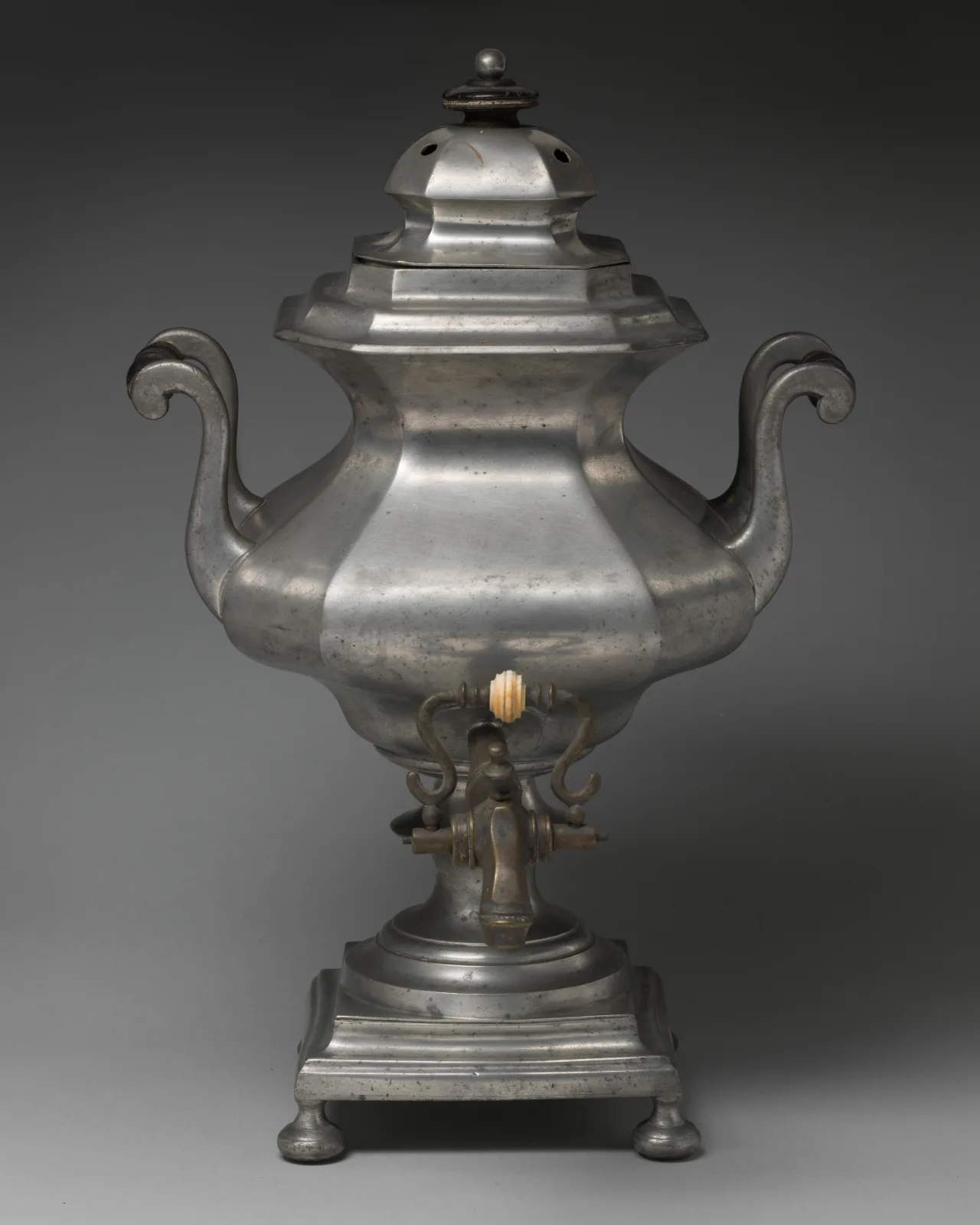 Tall grey Britannia metal tea urn with an octagonal body, two scrolled handles, a stepped lid, a tap with an ivory-coloured handle and a square stepped base