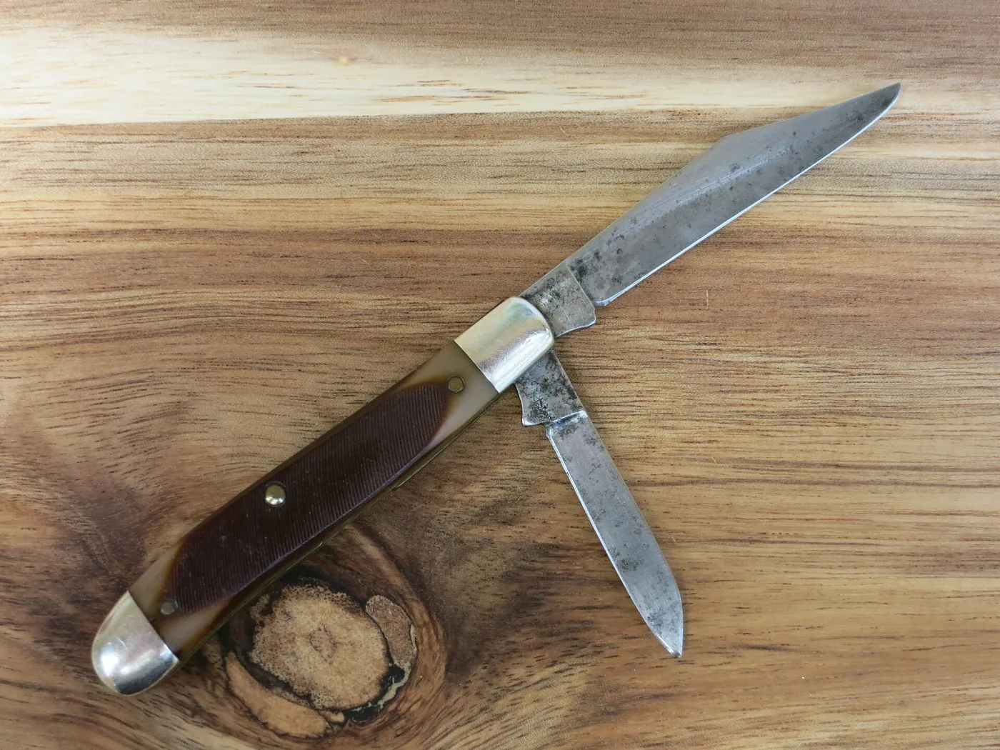 A U.S.-made Old Timer stockman on a wooden surface with its large clip blade and small spey blade open, both blades spotted and darkened with age, brown sawcut handles and nickel silver bolsters
