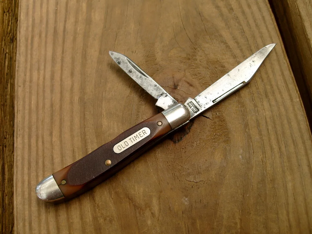 A small two-blade Old Timer peanut pocket knife on a wooden board with both blades open and brown sawcut handles, the tang stamp visible at the base of the main blade