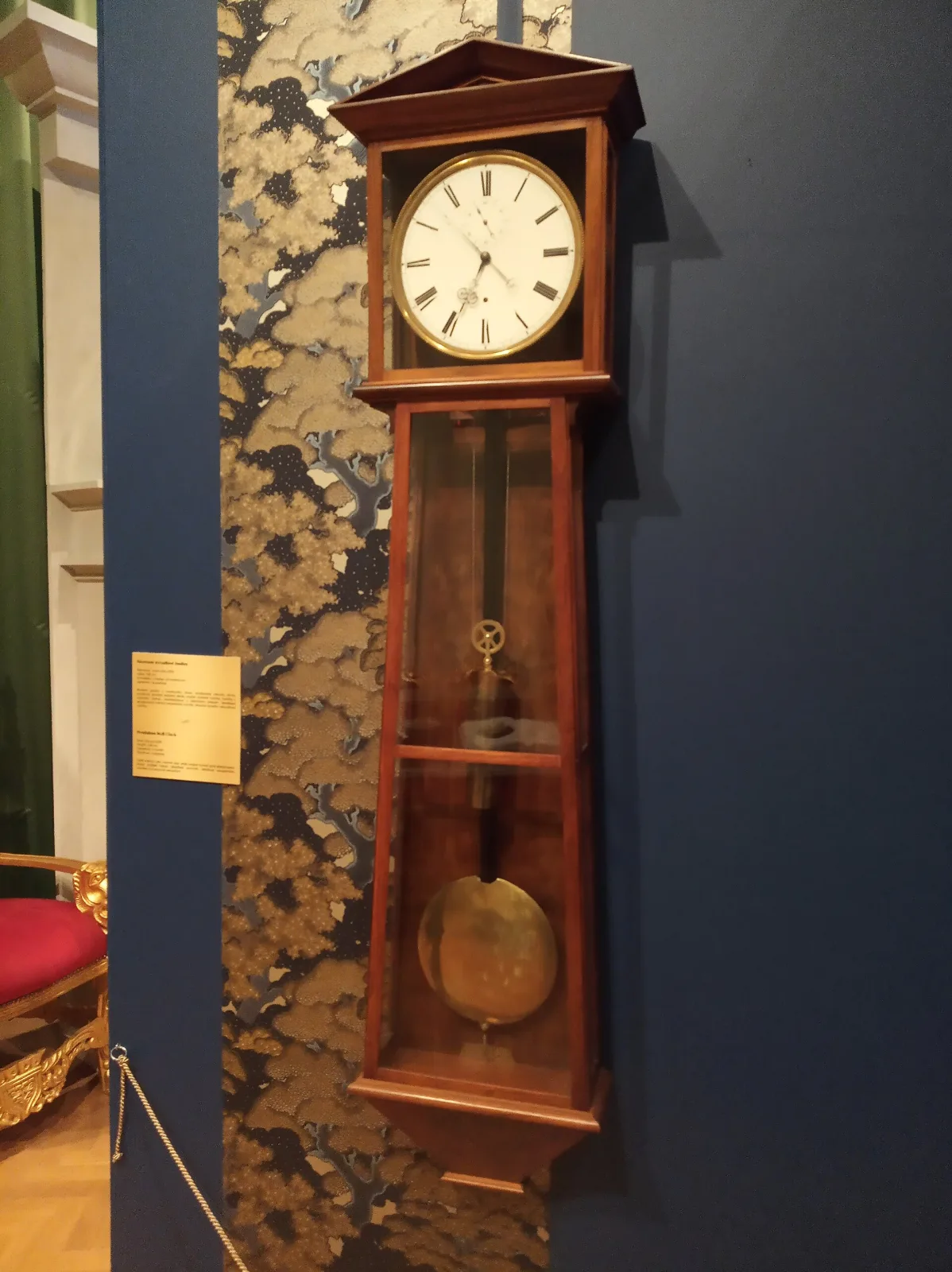 Plain walnut wall clock with a pediment top, a white enamel dial in a glazed head and a glazed trunk widening towards the base, with a single weight and a brass pendulum bob inside