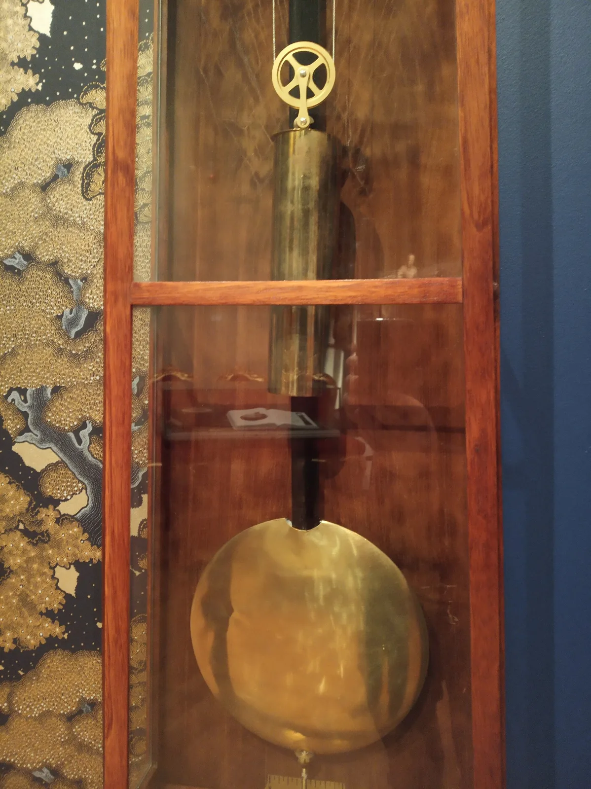 View through the glass of a walnut wall clock showing a single brass-cased cylindrical weight hanging from a small pulley wheel and, below it, the large brass bob of the pendulum on a dark wooden rod