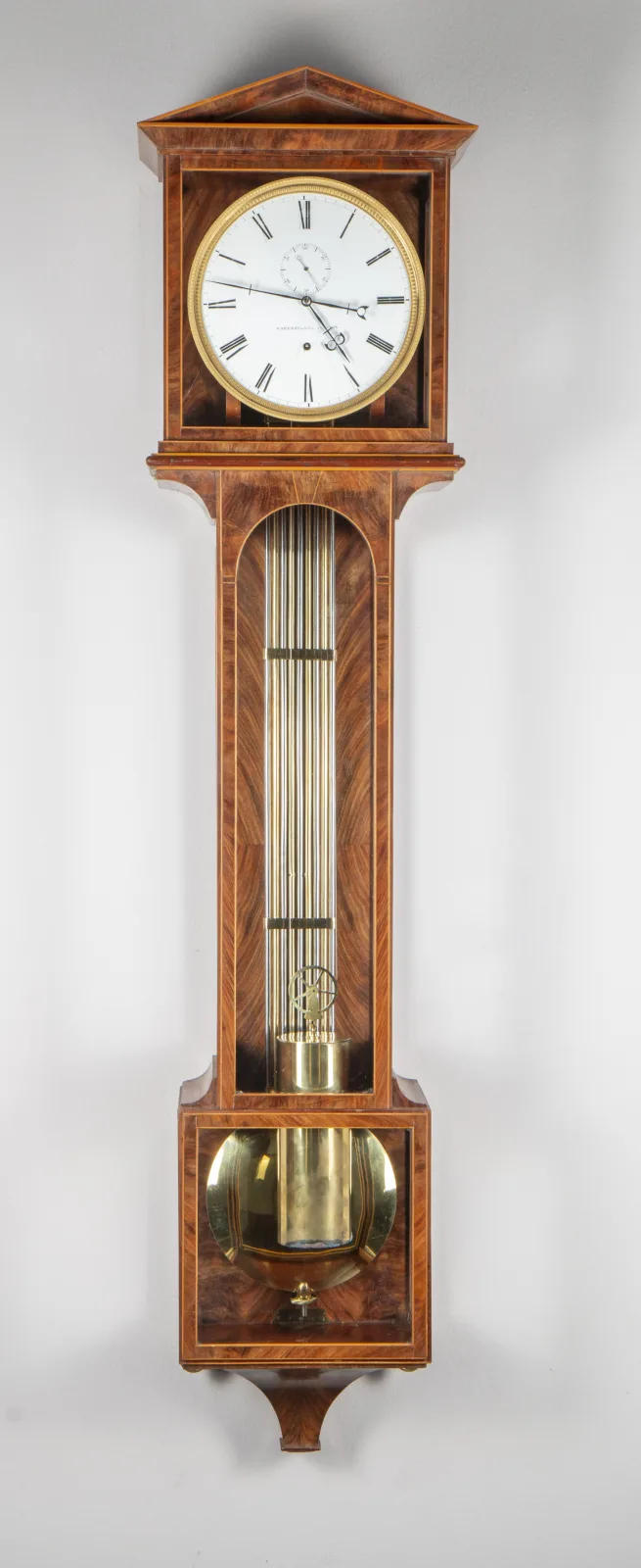 Slim wall regulator in figured veneer with a low triangular pediment, a white enamel dial with seconds subsidiary, a long glazed trunk showing a pendulum of many parallel rods and a brass bob in the lower glazed box
