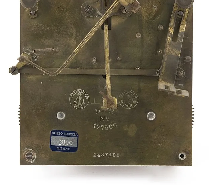 Brass back plate of a clock movement stamped with a round Gustav Becker Freiburg trademark showing a crowned anchor between the letters G and B, a round Medaille d'Or stamp, D.R.P. No 177600, a museum inventory sticker and the serial number 2437421