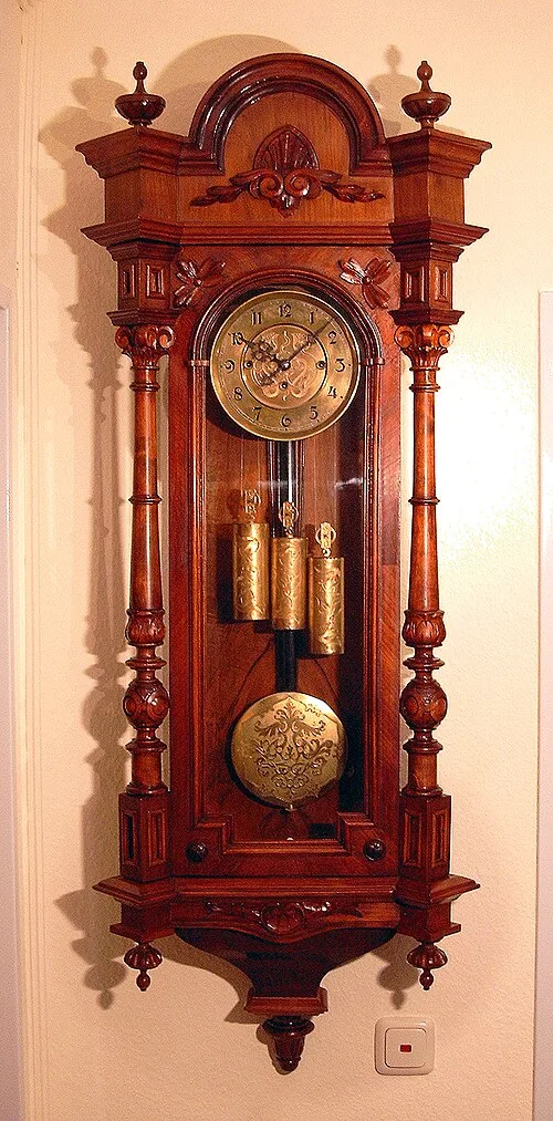 Wall clock in a red-brown carved case with turned columns ending in finials, an arched crest, a brass dial with a decorated centre and three brass-cased weights above an embossed pendulum bob