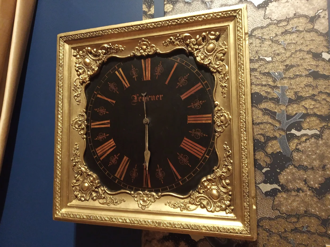 Square wall clock with a black dial painted with red Roman numerals and the maker's signature Lehrner, set in a heavily carved and gilded frame, photographed at an angle on a blue exhibition wall